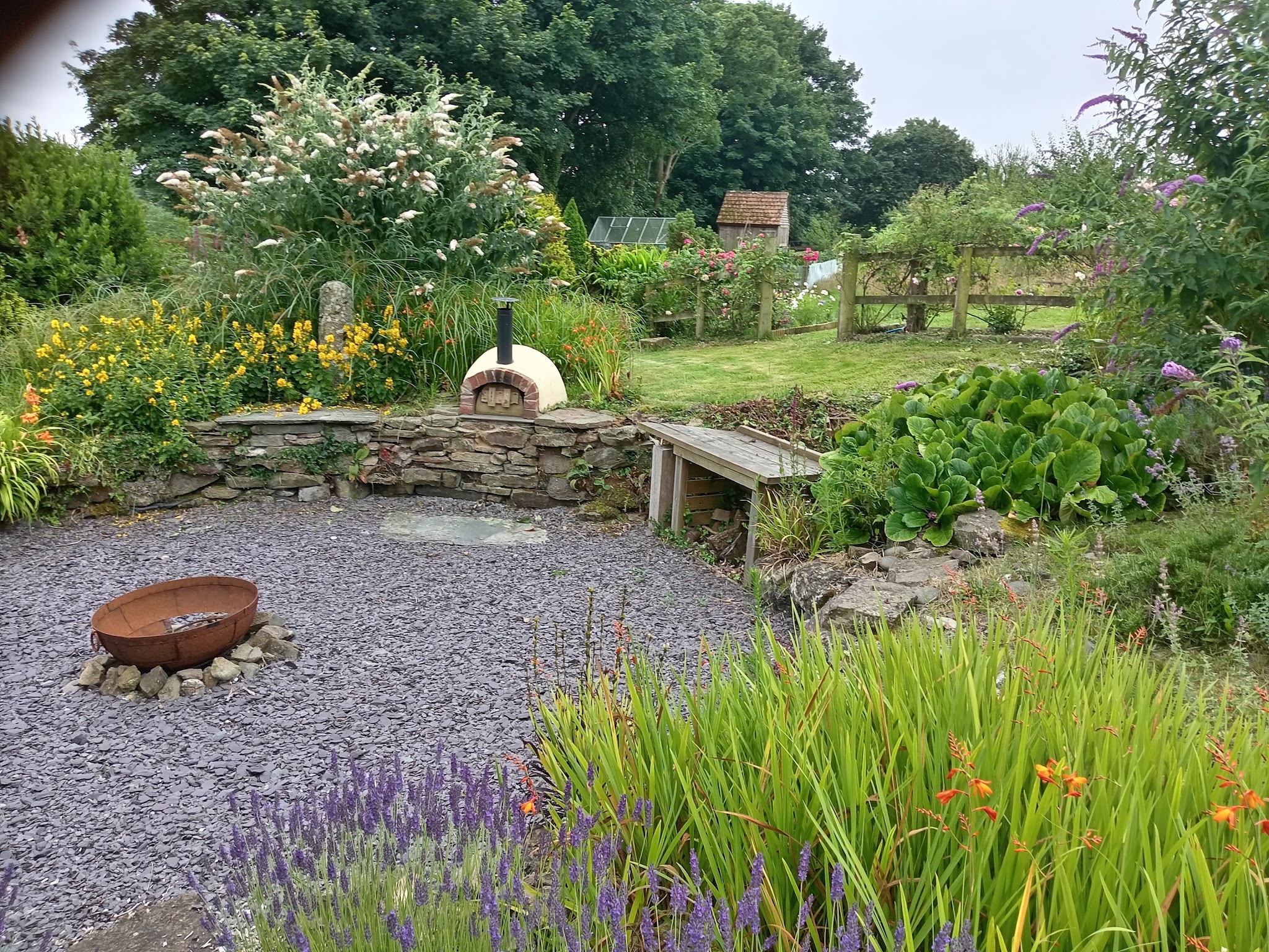 A slate gravel seating area with a fire bowl, low stone wall, lavender and a clay pizza oven in a lush cottage garden